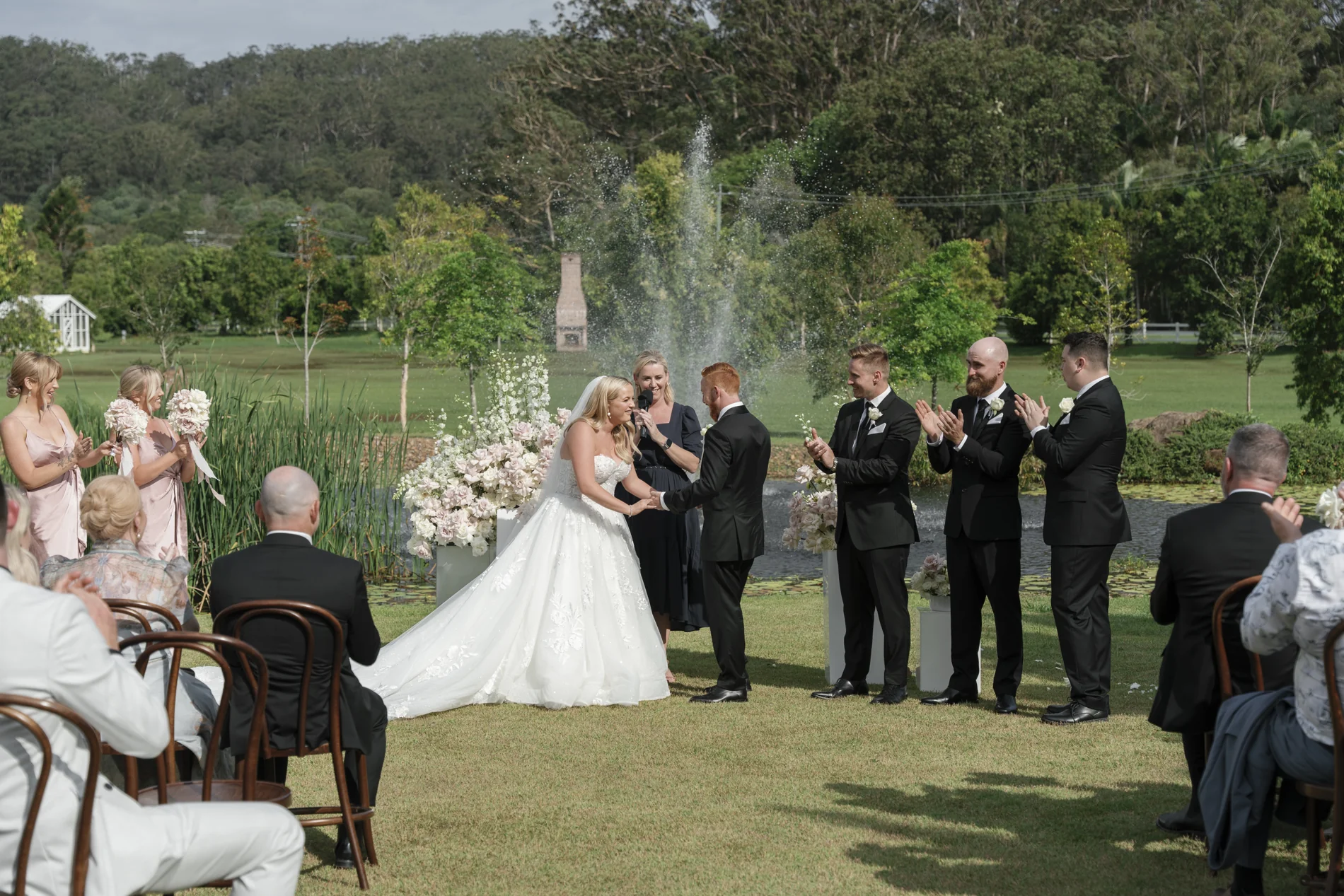 A wedding ceremony on a lawn with hills and a fountain beyond